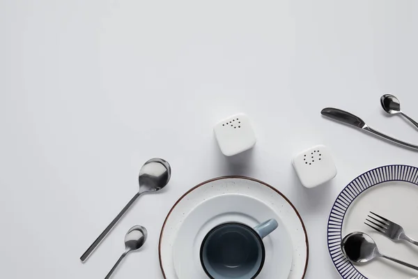 Vista superior de la bodega de sal, echador de pimienta, cucharas, cuchillo, tenedor, taza y platos en la mesa blanca - foto de stock
