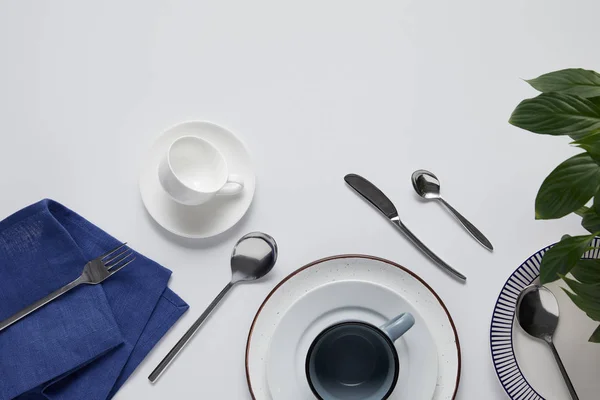 Top view of green leaves, various ceramic plates, cup, kitchen towel, fork, spoons and knife on white table — Stock Photo