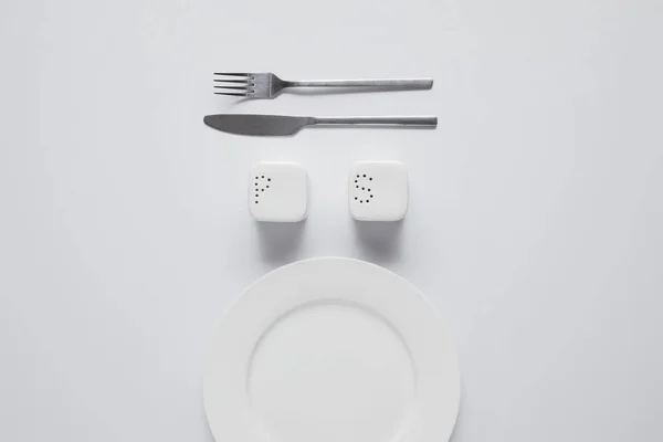 Draufsicht auf arrangierten Teller, Gabel, Messer, Salzkeller und Pfefferstreuer auf weißem Tisch, minimalistisches Konzept — Stockfoto