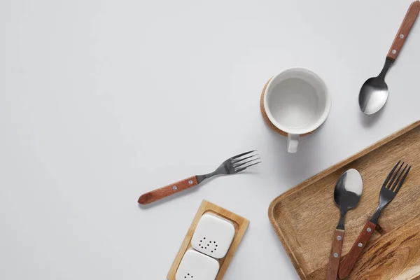 Top view of cup, spoons, forks, wooden tray, saltcellar and pepper caster on white table — Stock Photo