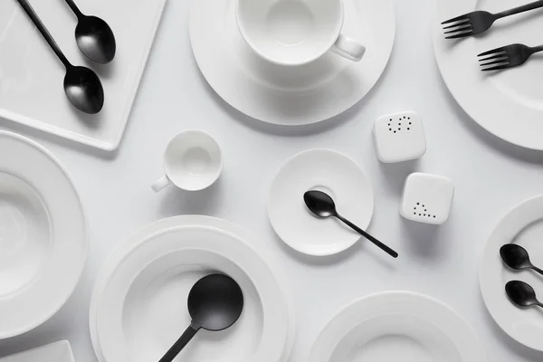 Top view of black spoons, forks, salad spoon, different plates, bowl, saltcellar, pepper caster and cup on white table — Stock Photo