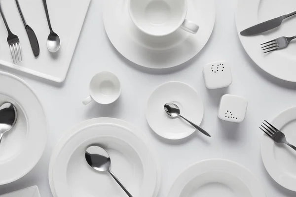 Vista dall'alto di cucchiai, forchette, coltelli, piatti diversi, ciotola, saliera, pepe caster e tazza sul tavolo bianco — Foto stock