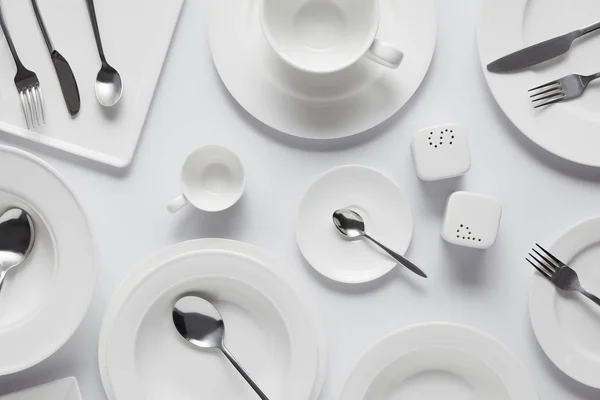 Top view of spoons, forks, knives, different plates, bowl, saltcellar, pepper caster and cup on white table — Stock Photo