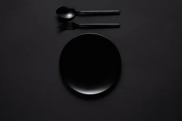 Vue de dessus de la plaque noire, fourchette et cuillère sur table noire, concept minimaliste — Photo de stock
