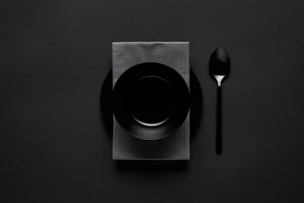 Top view of black bowl, spoon, kitchen towel and plate on black table, minimalistic concept — Stock Photo