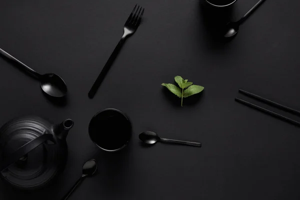 Vista superior do bule preto, pauzinhos, várias colheres, garfo, xícara e folhas de hortelã na mesa preta — Fotografia de Stock