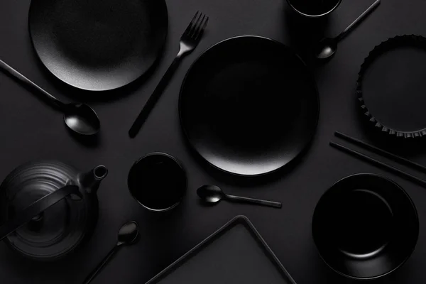 Vista dall'alto di teiera nera, piatti, ciotola, tazze, vassoio, cucchiai, bacchette, forchetta e teglia su tavolo nero — Foto stock