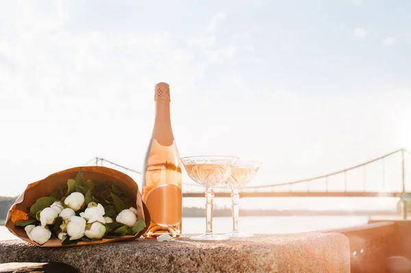 Close-up view of beautiful bouquet of flowers, two glasses and bottle of champagne near river at sunset — Stock Photo