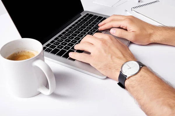 Tiro recortado de homem digitando com laptop enquanto trabalha com xícara de café em primeiro plano na superfície branca — Fotografia de Stock