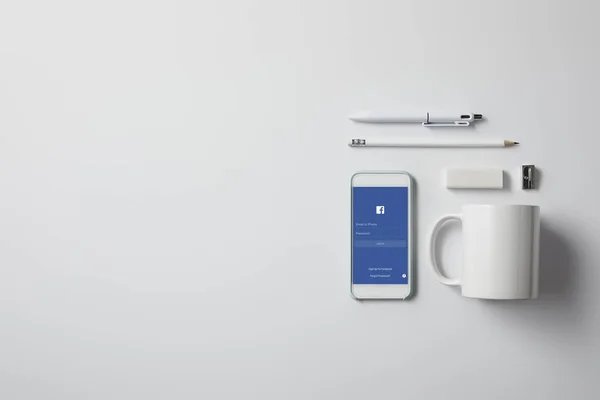 Top view of smartphone with facebook app and various supplies on white tabletop — Stock Photo