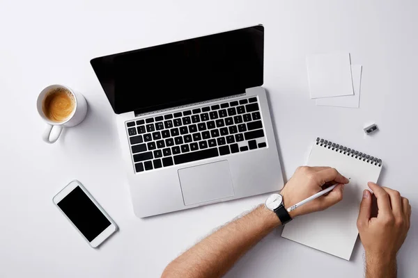 Cropped shot of man writing in notebook at workplace with gadgets and coffee cup on white surface for mockup — Stock Photo
