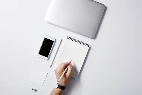 Tiro recortado de homem escrevendo em notebook no local de trabalho com gadgets na superfície branca para maquete — Fotografia de Stock
