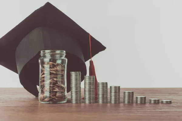 Stacks of coins, jar with coins and graduation cap on wooden table, saving concept — Stock Photo