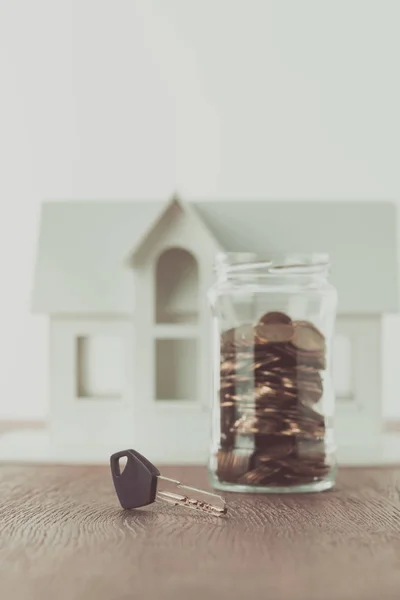 Chave perto de frasco de moedas em mesa com pequena casa no fundo, economizando conceito — Fotografia de Stock