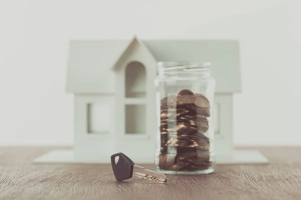 Chave perto de frasco de moedas na mesa com pequena casa no fundo, economizando conceito — Fotografia de Stock