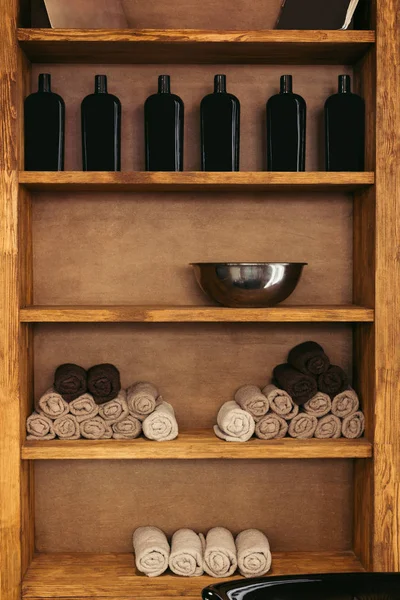 Cuenco de metal vacío, toallas enrolladas y botellas de vidrio en estantes de madera en la barbería - foto de stock