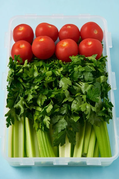Primer plano vista del recipiente de alimentos lleno de tomates frescos, perejil y apio sobre fondo azul - foto de stock