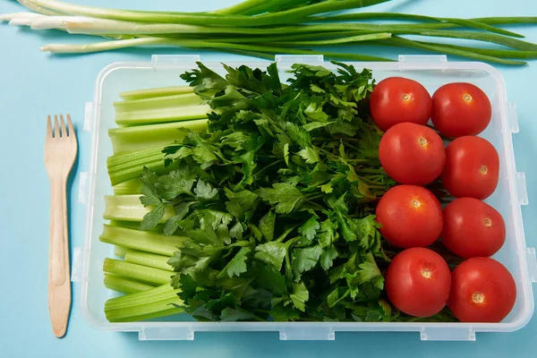 Primer plano vista de tenedor y contenedor de alimentos lleno de tomates frescos, perejil y apio sobre fondo azul - foto de stock