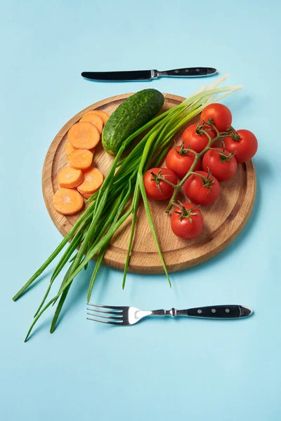 Vista de cerca de cubiertos y verduras frescas surtidos en la tabla de cortar de madera sobre fondo azul - foto de stock