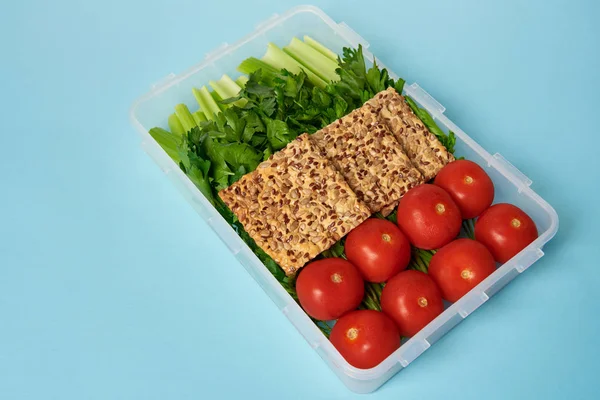 Vista de perto do recipiente de alimentos cheio de legumes saudáveis e biscoitos com sementes no fundo azul — Fotografia de Stock