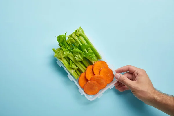 Plan recadré d'une main masculine et d'un récipient rempli de tranches de céleri et de carotte isolées sur du bleu — Photo de stock