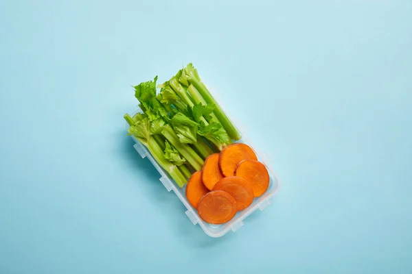 Vista superior del recipiente lleno de apio sano y rodajas de zanahoria aisladas en azul - foto de stock