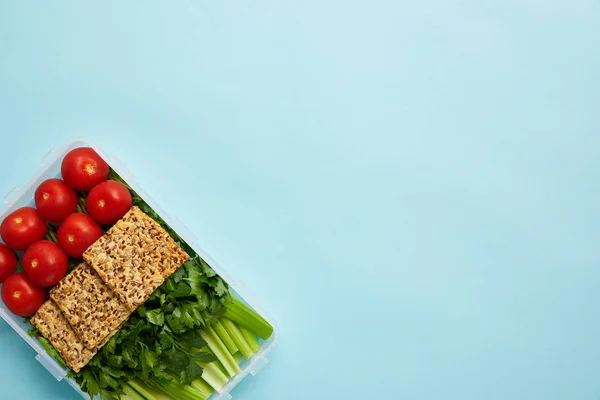 Vista superior del contenedor de alimentos lleno de comida saludable con verduras y galletas aisladas en azul - foto de stock