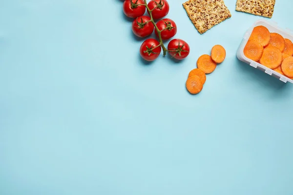 Top view of arrangement of food container with fresh carrot slices, tomatoes and cookies isolated on blue — Stock Photo