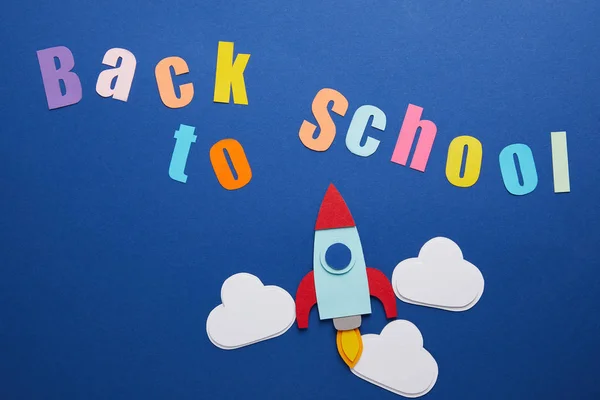 Vue du dessus du lettrage de retour à l'école avec nuages et fusée sur fond bleu — Stock Photo