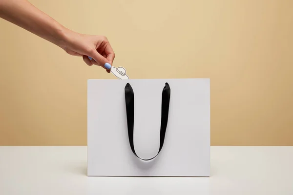 Imagen recortada de la mujer poniendo sombrero de papel en la bolsa de compras aislado en beige - foto de stock