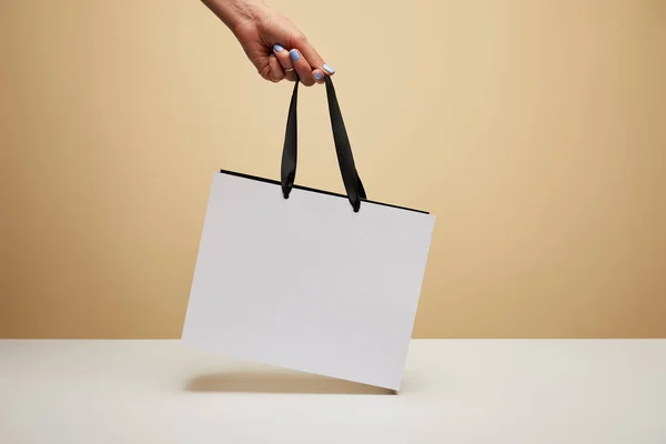 Cropped image of woman holding white shopping bag above white table isolated on beige — Stock Photo