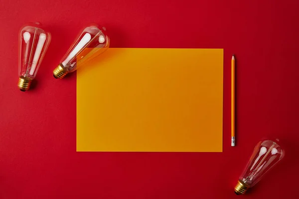 Vista superior de papel amarelo em branco com lápis e lâmpadas incandescentes na superfície vermelha — Fotografia de Stock