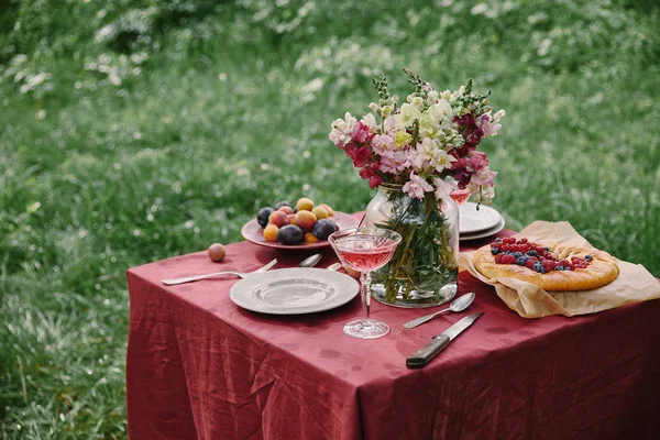Copa de vino, tarta de bayas y ramo de flores en la mesa en el jardín verde - foto de stock