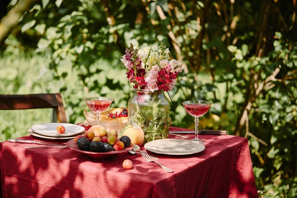 Fruits, wineglasses and bouquet of flowers on table in garden — Stock Photo