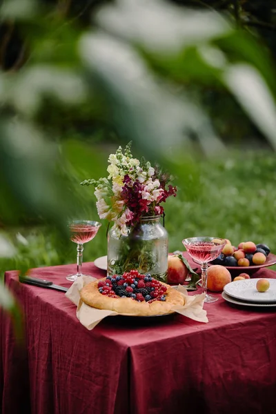 Bouquet of flowers in glass jar, berries pie and wineglasses on table in garden — Stock Photo