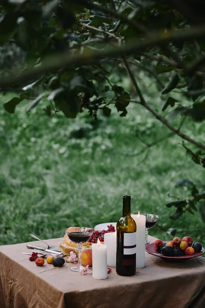 Velas, botella de vino y frutas en la mesa en el jardín para la cena - foto de stock