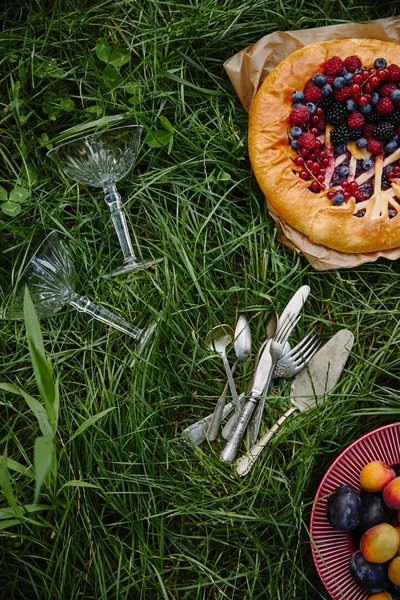 Vista superior del pastel con bayas, plato con frutas y utensilios sobre hierba verde - foto de stock