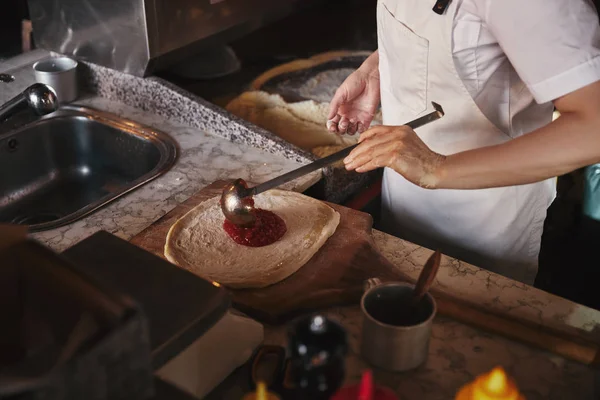 Plan recadré de cuisinier verser du ketchup sur la pâte à pizza à la cuisine du restaurant — Photo de stock