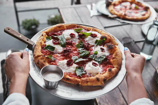 Colpo ritagliato di donna che trasporta piatto con deliziosa pizza da servire sul tavolo al ristorante — Foto stock