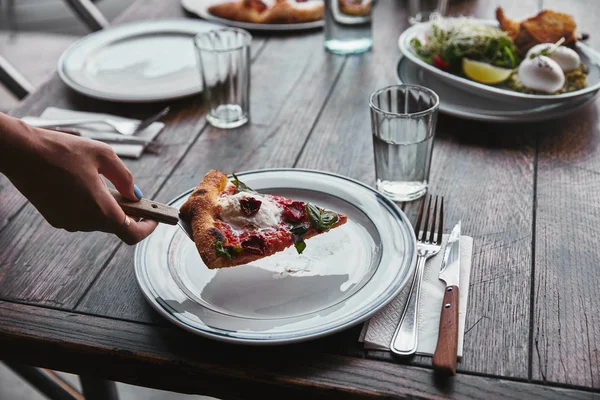 Обрезанный снимок женщины, подающей кусочек вкусной пиццы на тарелке в ресторане — стоковое фото