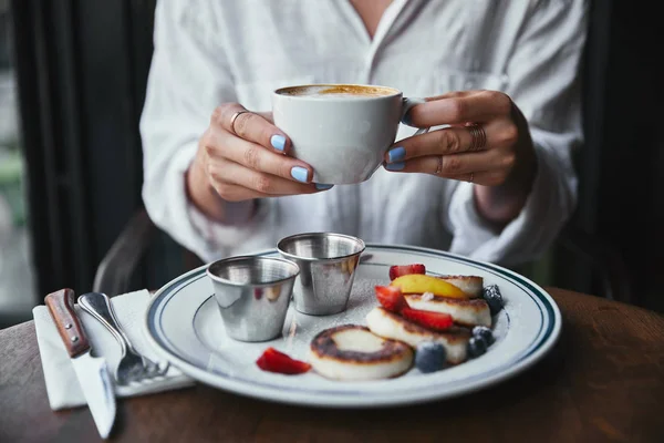 Tiro cortado de mulher segurando xícara de café no restaurante acima panquecas de queijo na mesa — Fotografia de Stock