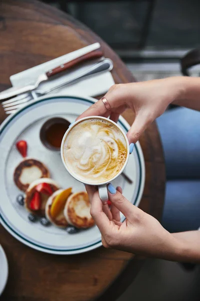 Cropped shot of woman holding cup of coffee above syrniki on table at restaurant — Stock Photo