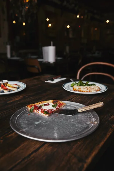 Slice of pizza with server on metal tray and wooden table with plates of lasagne and syrniki — Stock Photo