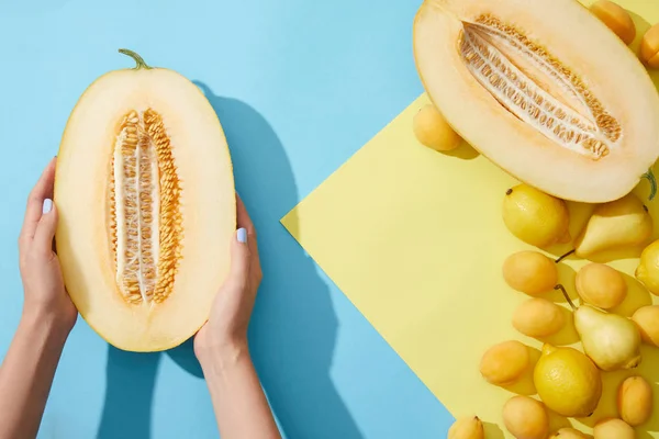 Vista dall'alto parziale della persona che tiene in mano melone dimezzato e pere mature fresche, albicocche e limoni su giallo e blu — Foto stock