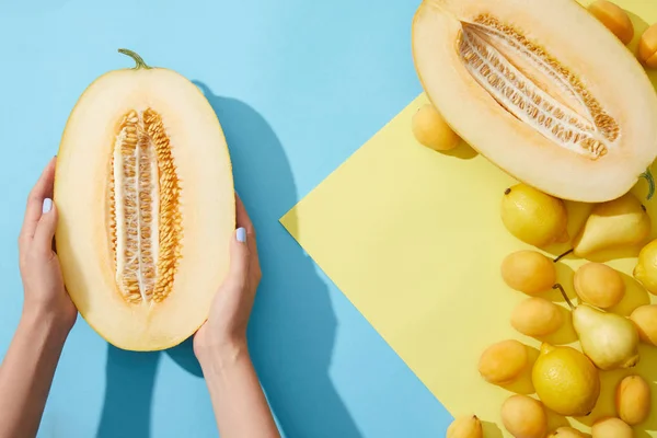 Partial top view of person holding halved melon in hands and fresh ripe pears, apricots and lemons on yellow and blue — Stock Photo