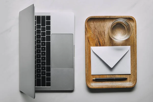 Arbeitsplatzvorlage mit Laptop und Holztablett mit Umschlag und Wasser auf weißem Marmorhintergrund — Stockfoto