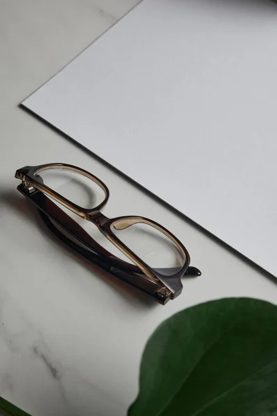 Очки и чистая бумага на фоне белого мрамора с зеленым листом — стоковое фото