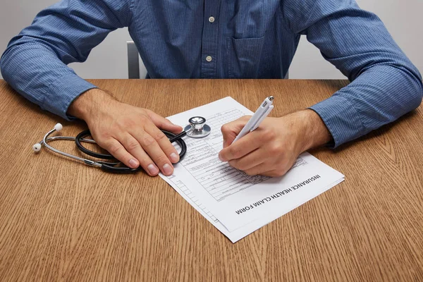 Tiro recortado de la persona que sostiene estetoscopio y firmar el formulario de seguro en la mesa de madera — Stock Photo