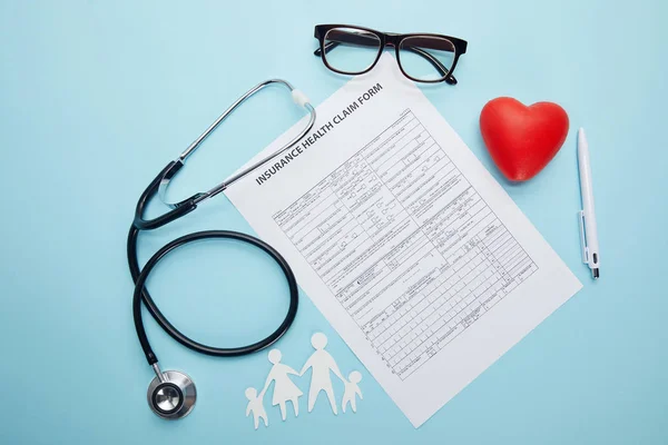 Vue du dessus du formulaire de demande de règlement d'assurance maladie, des lunettes, de la famille coupée en papier, du symbole du coeur rouge et du stéthoscope sur bleu — Photo de stock