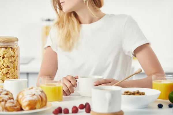 Vista parcial da mulher sentada à mesa com croissants, xícaras de café e copos de suco para o café da manhã — Fotografia de Stock